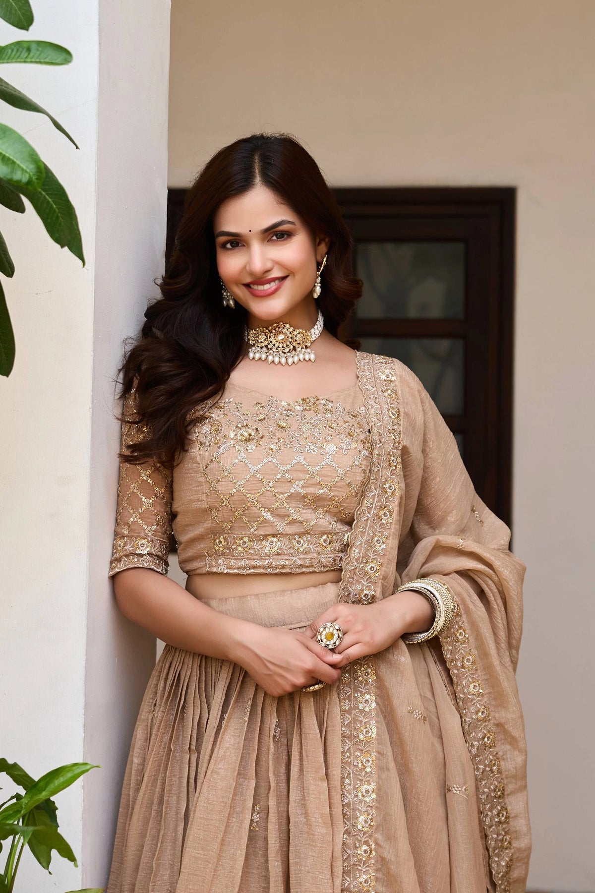 Woman in a beige embroidered outfit standing indoors with plants in the background