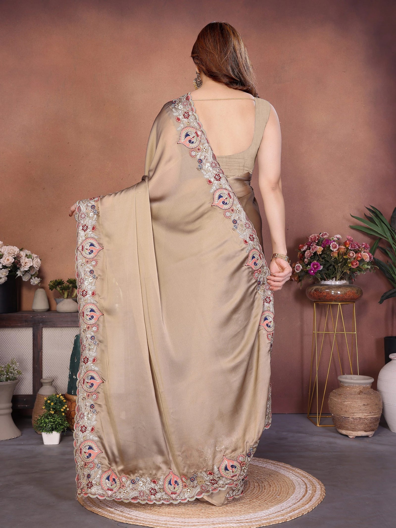 Woman wearing a beige saree with floral embroidery, standing in a room with decorative plants and flowers.