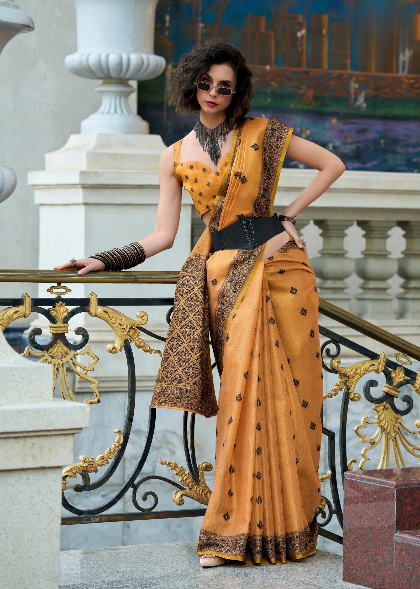 Woman in a mustard yellow saree with black patterns standing on a decorative staircase.