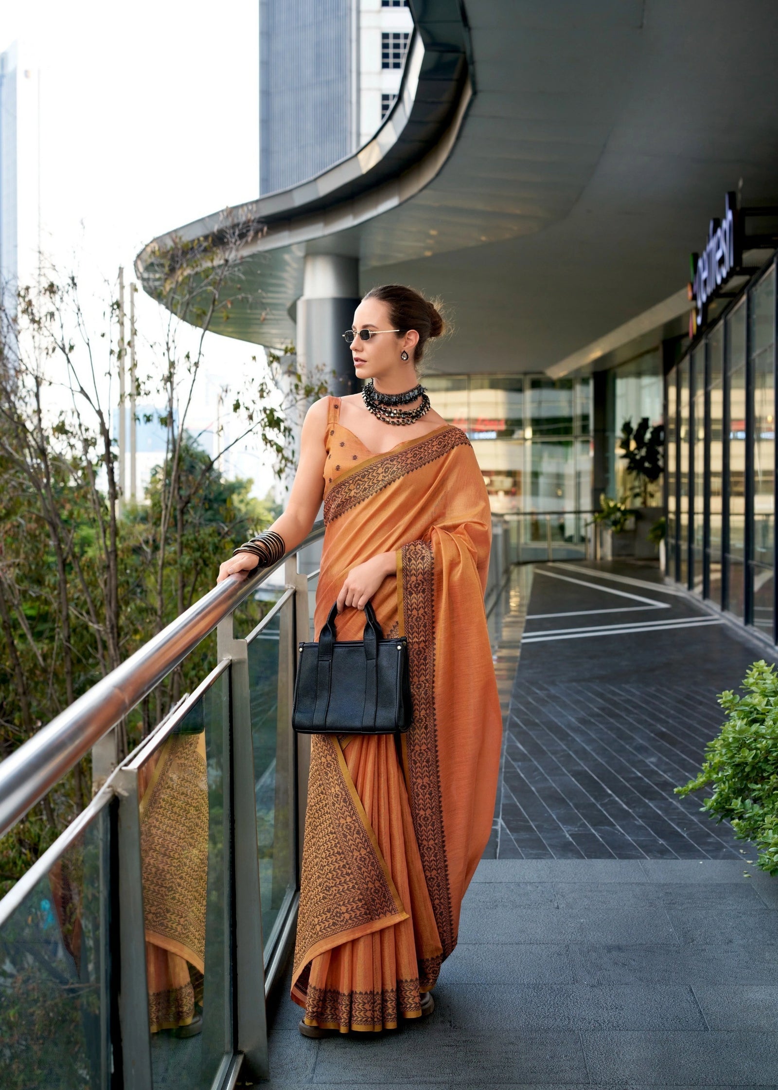 Woman in an orange saree standing on a balcony with modern architecture in the background