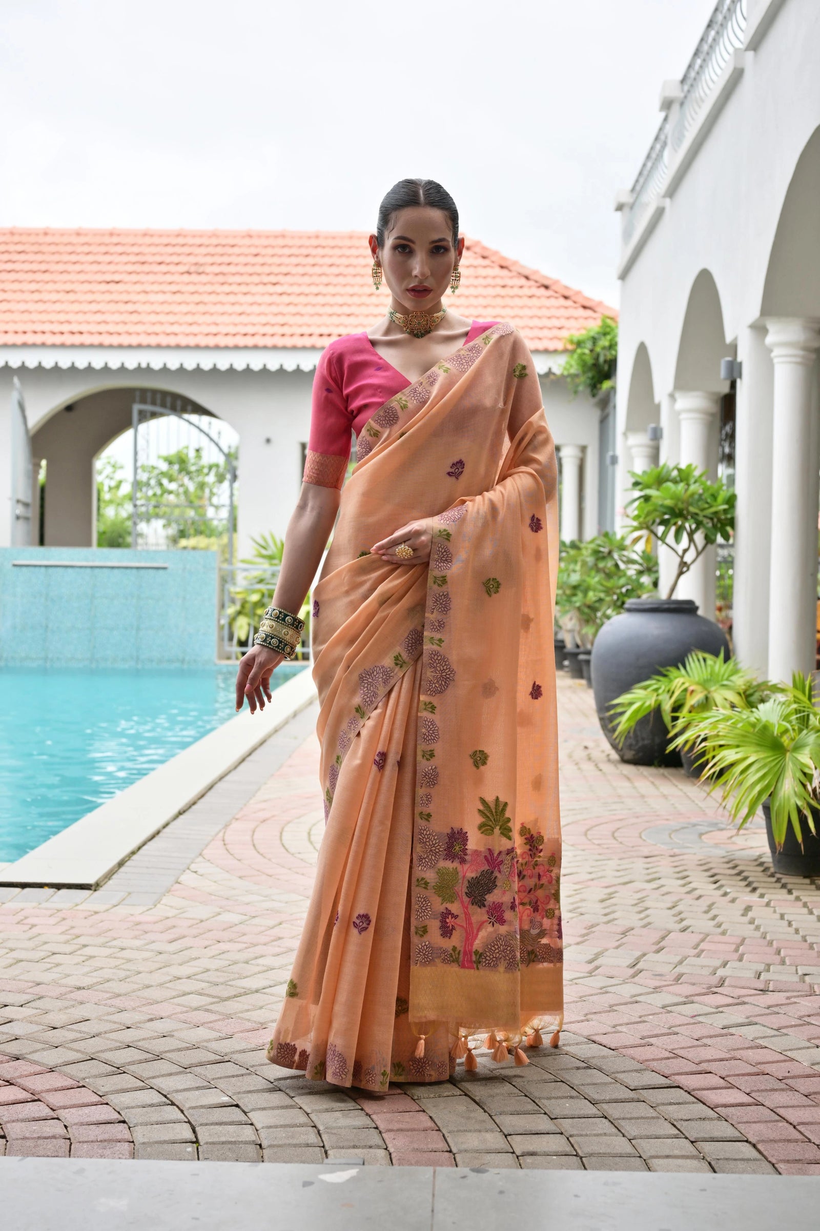 Woman in a peach saree with floral patterns standing by a poolside.