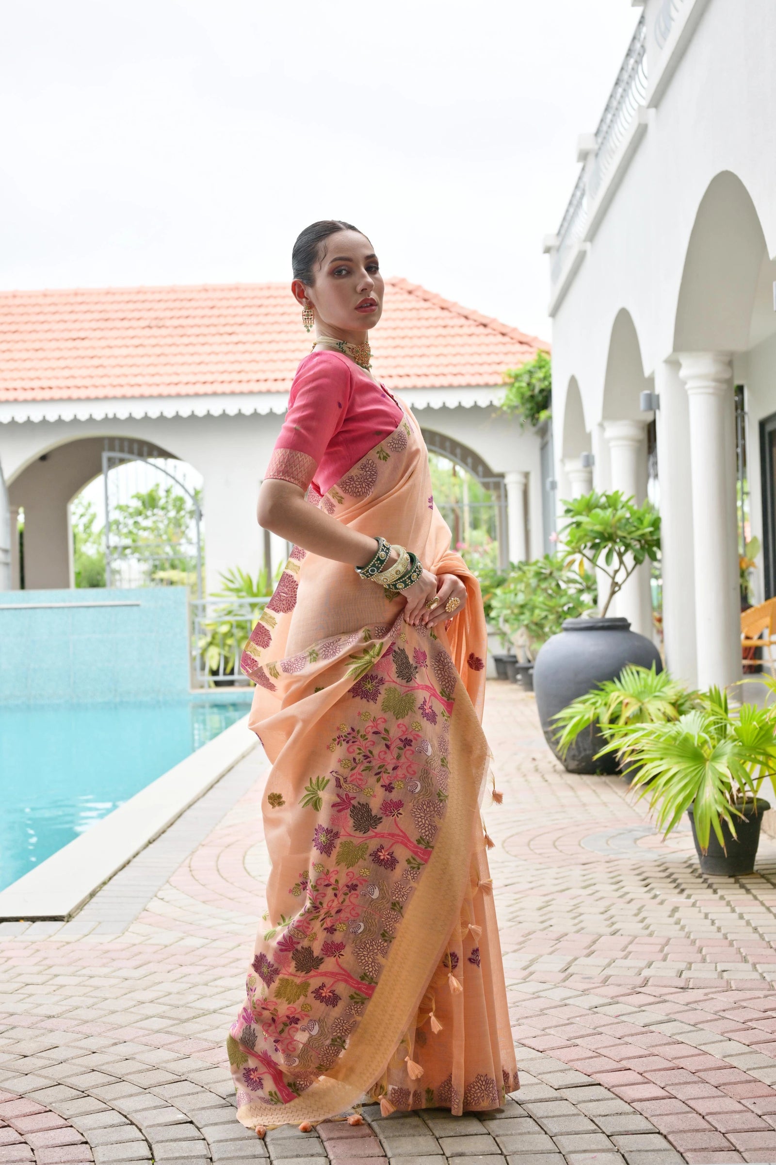 Woman in a floral saree standing by a poolside with a white building and greenery in the background.