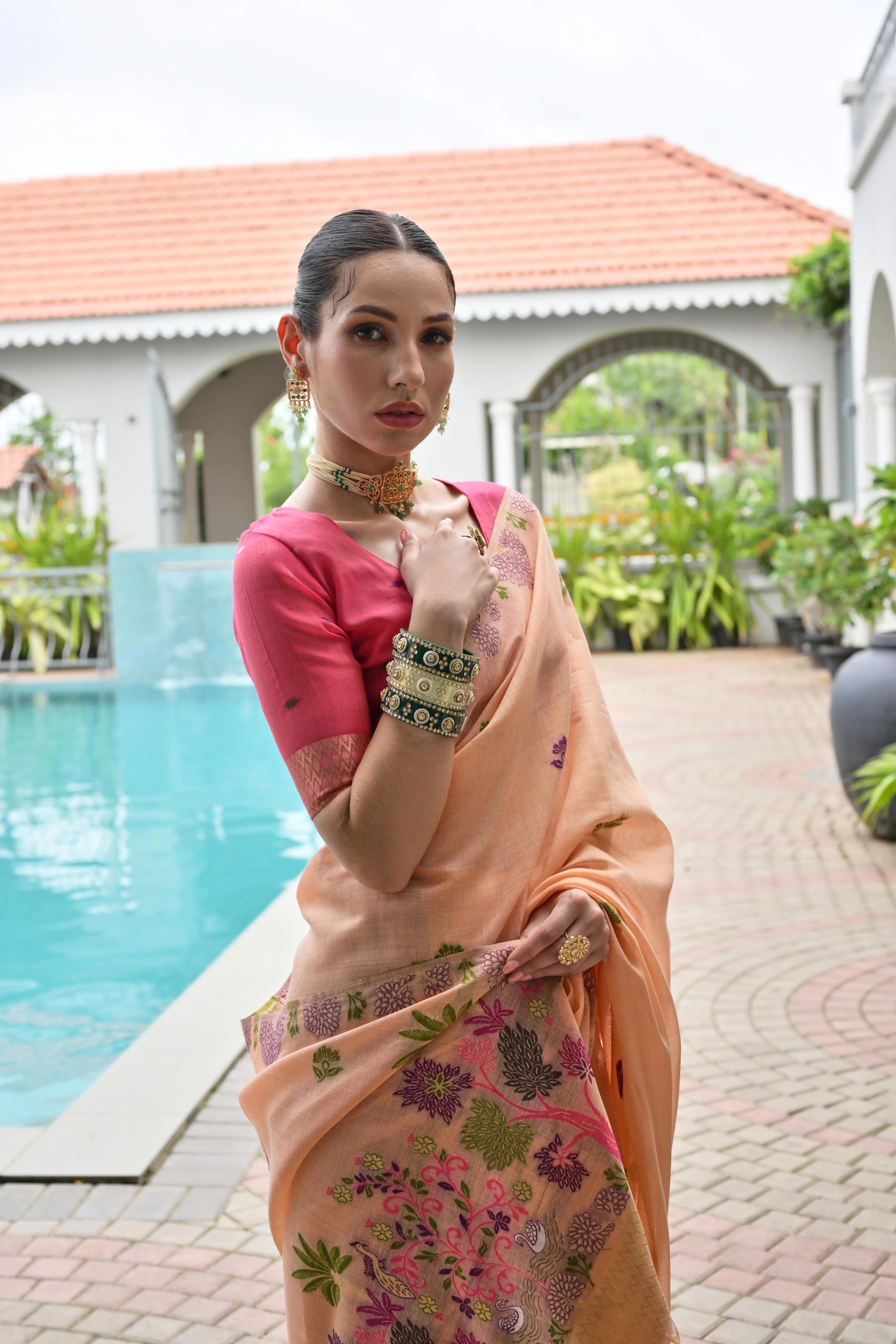 Woman in a pink blouse and peach saree with floral patterns by a poolside.