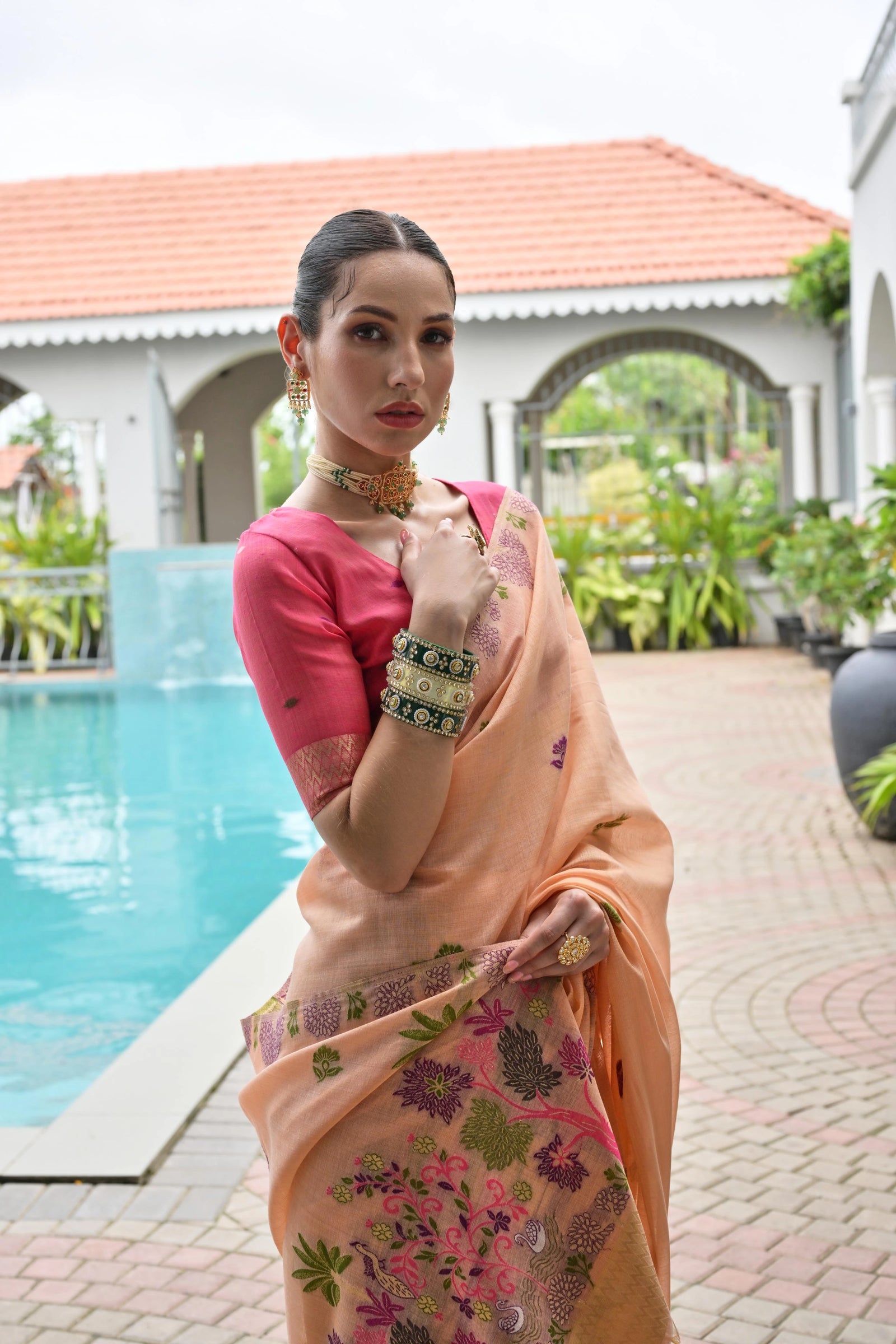 Woman in a pink blouse and peach saree with floral patterns by a poolside.