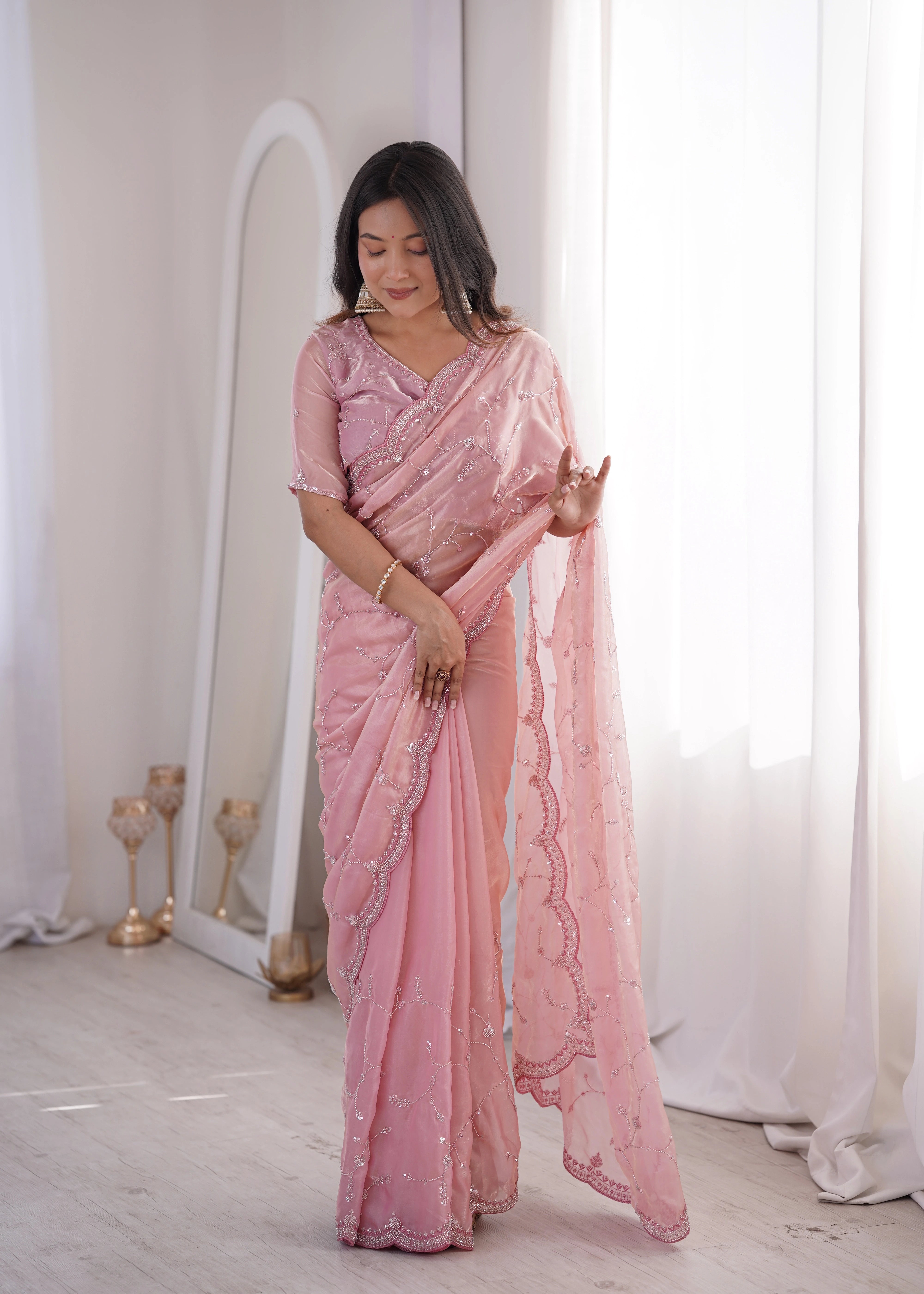 Woman wearing a pink saree in a room with white curtains and a mirror.