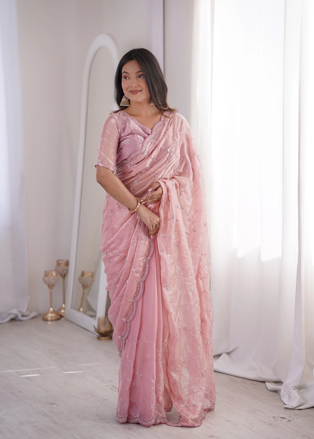 Woman wearing a pink saree in a minimalistic setting with white curtains and a white floor.