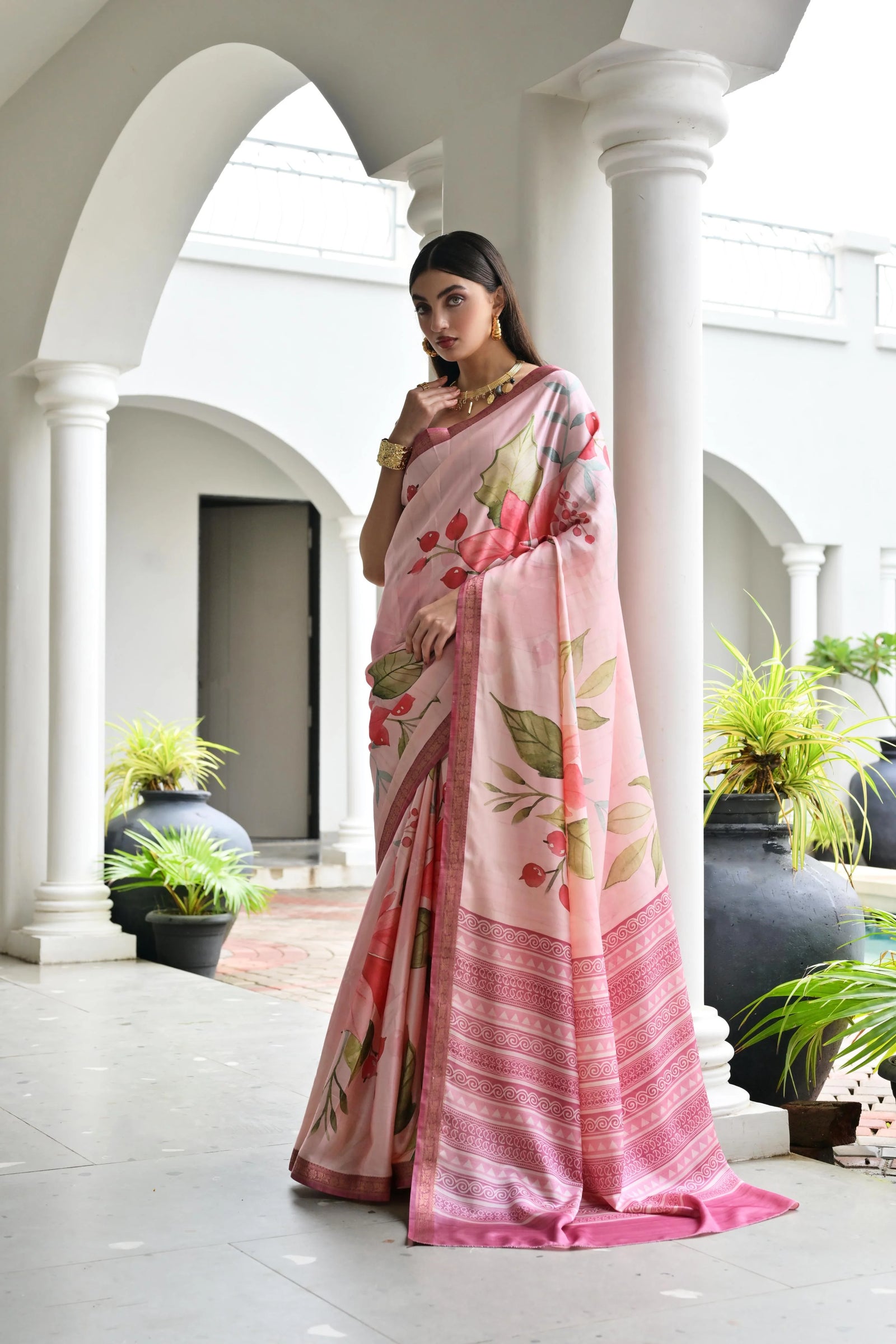 Woman in a pink saree with floral patterns standing in an elegant outdoor setting.