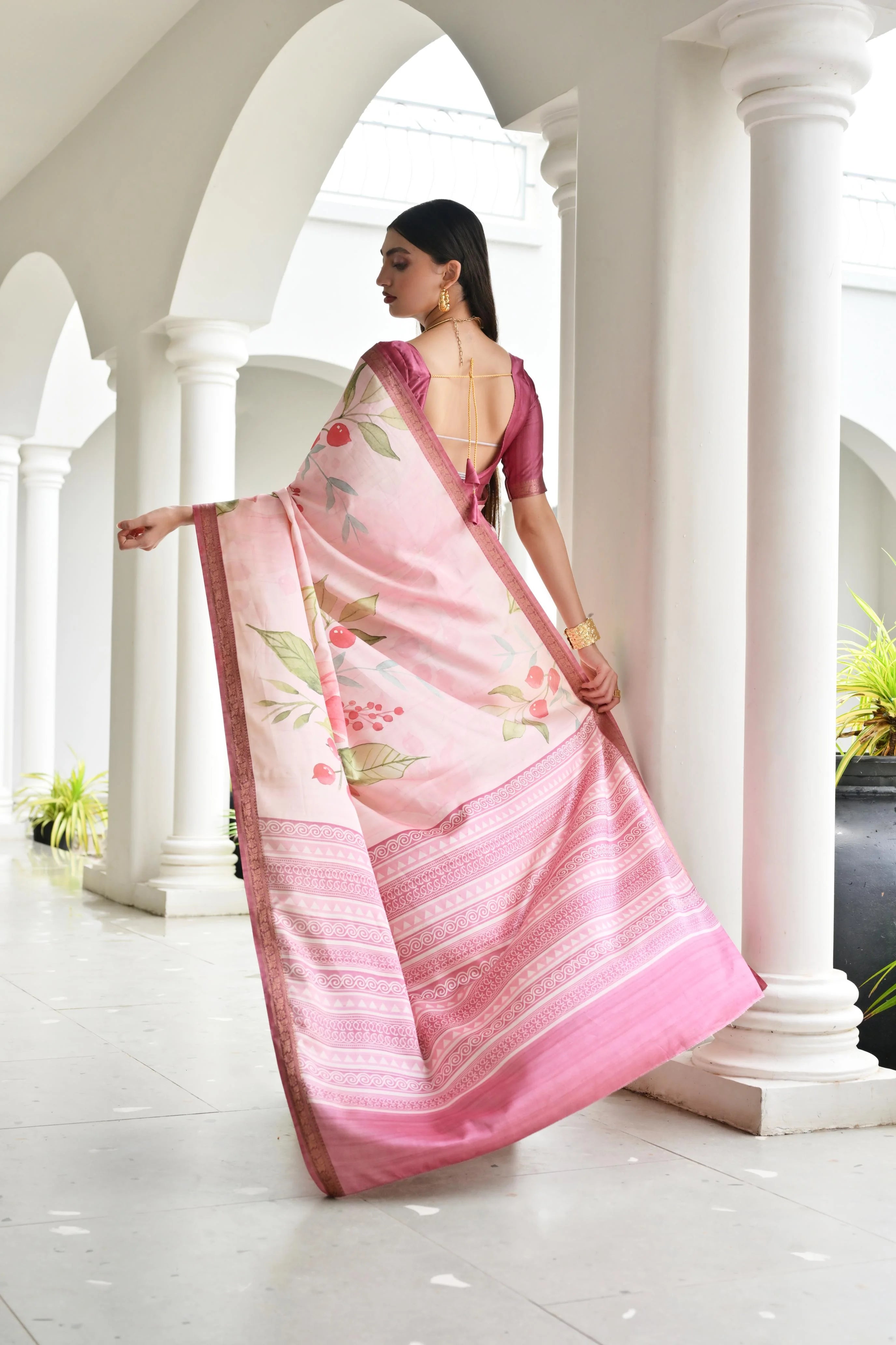 Woman in a pink saree with floral patterns standing in a white architectural setting.