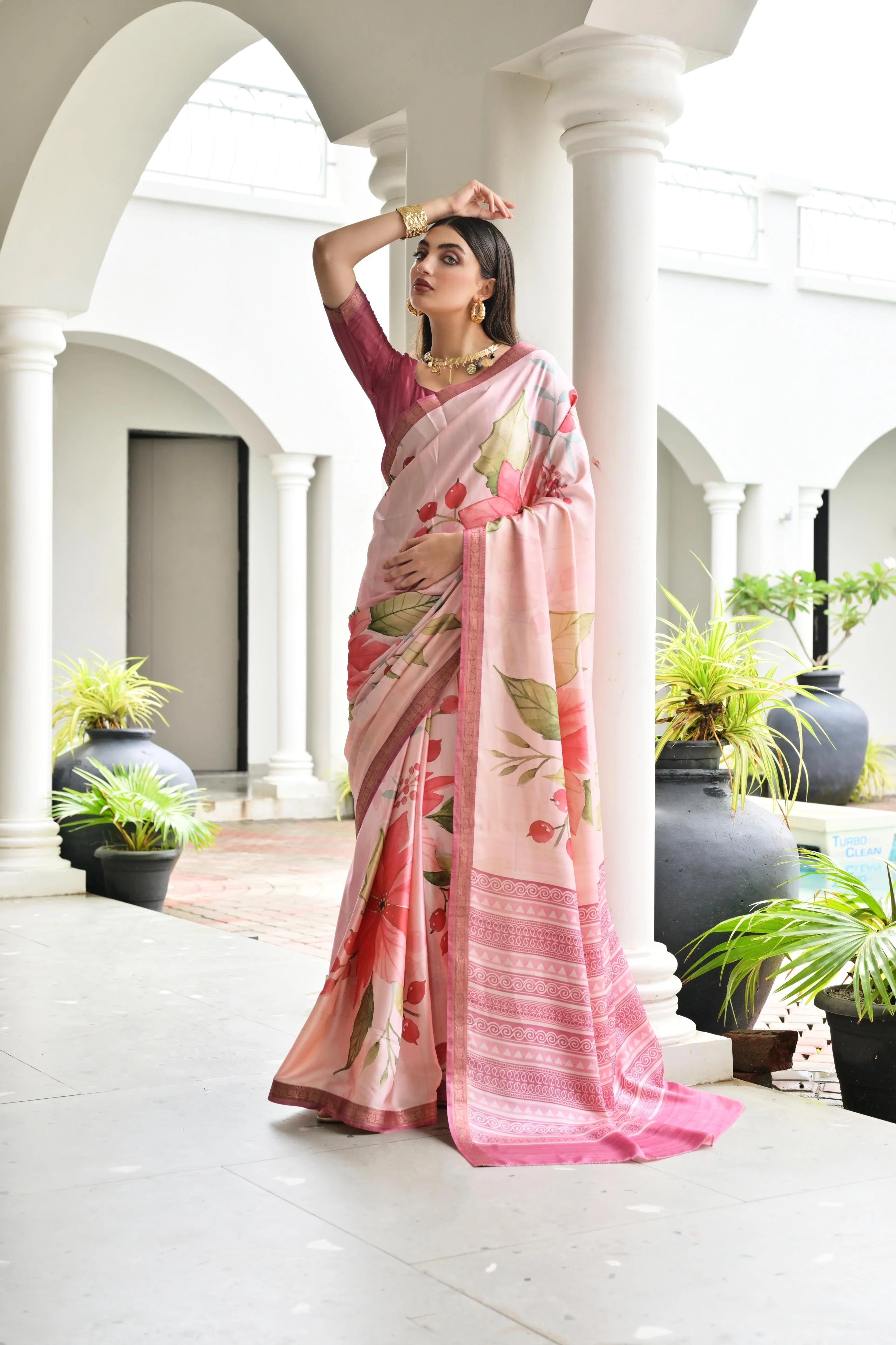 Woman in a pink saree with floral patterns standing in an elegant outdoor setting.