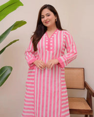 Woman wearing a pink and white striped dress standing next to a plant and wooden chair.