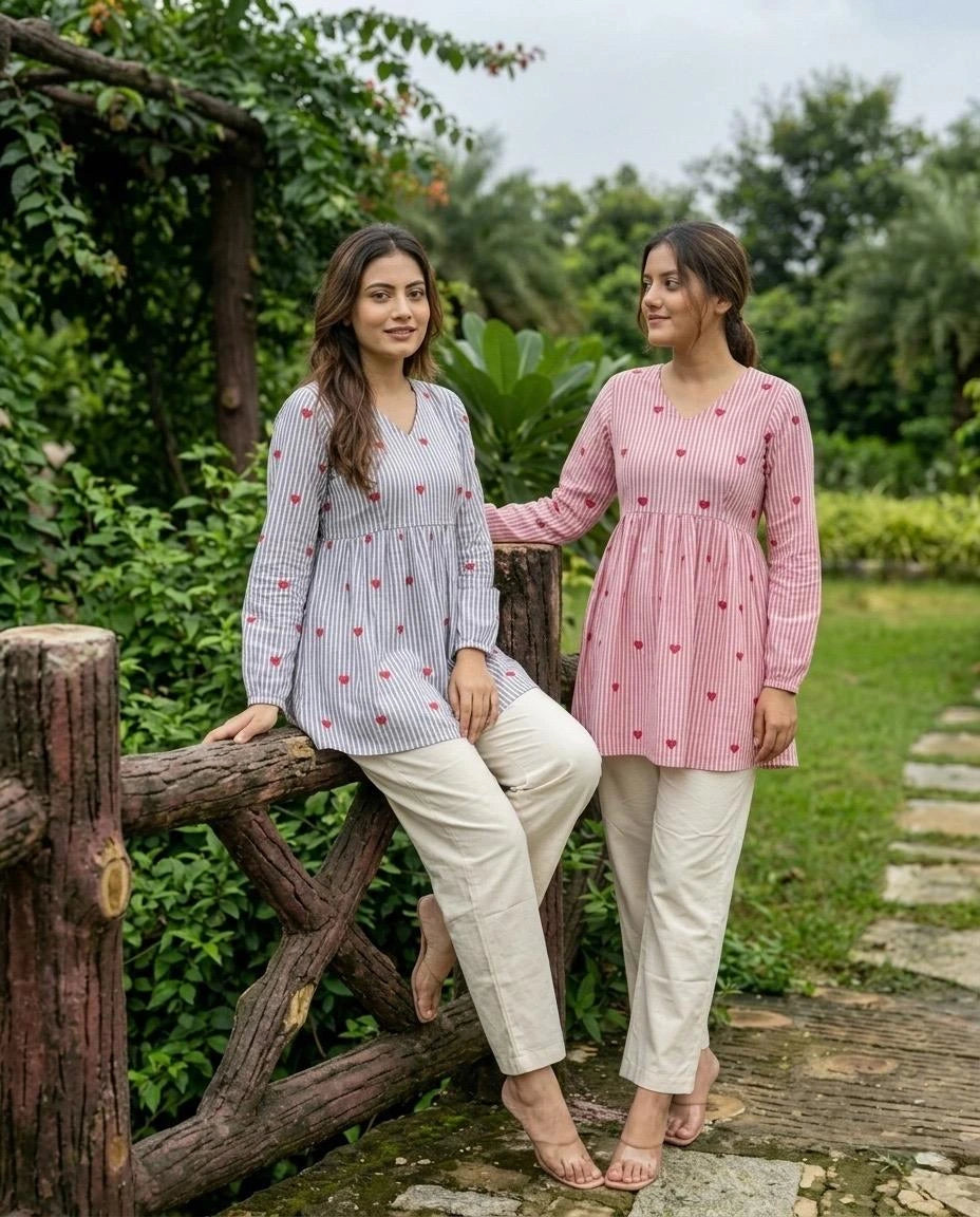 Two women in patterned tops and white pants sitting on a wooden fence outdoors.
