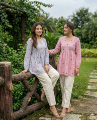 Two women in patterned tops and white pants sitting on a wooden fence outdoors.