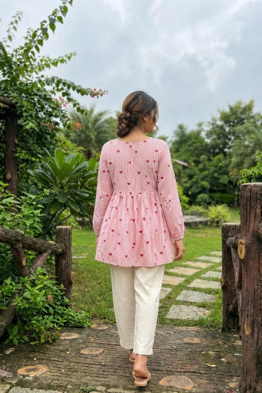 Woman in a pink checkered top and white pants walking through a garden.
