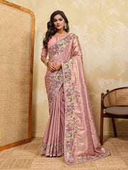 Woman wearing a pink saree with floral patterns in a room with a textured wall and chair.