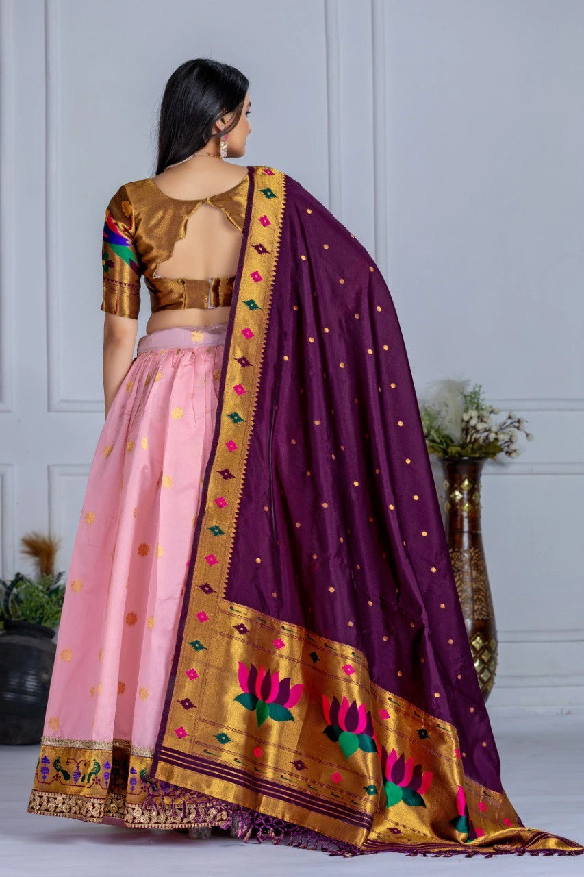 Woman wearing a traditional outfit with a purple dupatta and pink skirt against a white wall.