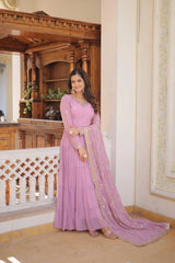 Woman in a pink saree standing in an ornate room with wooden furniture.