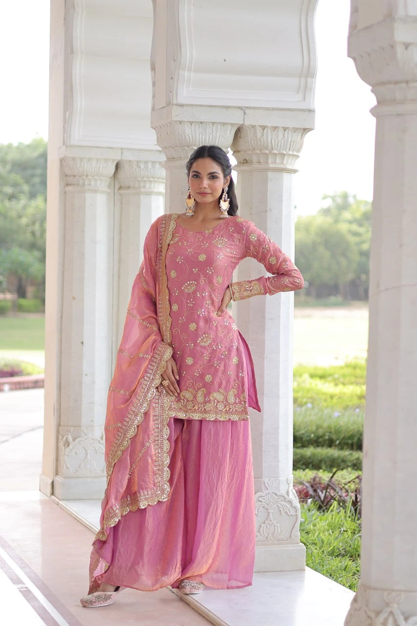 Woman in a pink traditional outfit standing in front of white architectural columns with greenery in the background.
