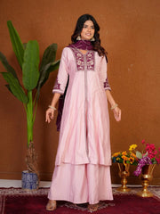 Woman wearing a pink traditional outfit with intricate embroidery, standing against a brown wall with decorative elements.