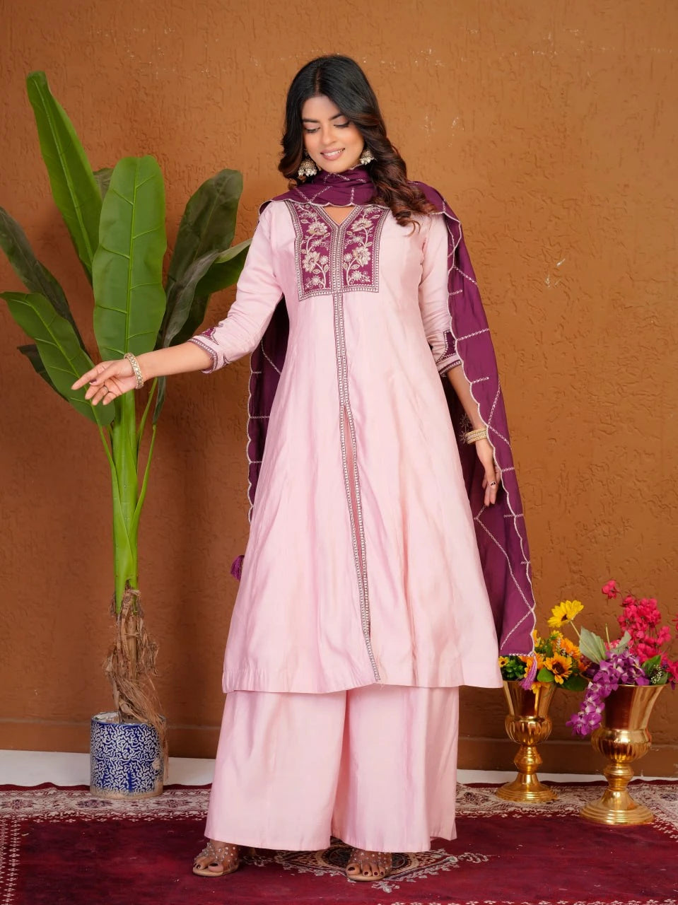 Woman in a pink traditional outfit with purple accents standing against a brown wall with decorative elements.