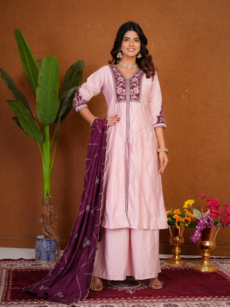 Woman in a pink and purple traditional outfit standing against a brown wall with decorative elements.