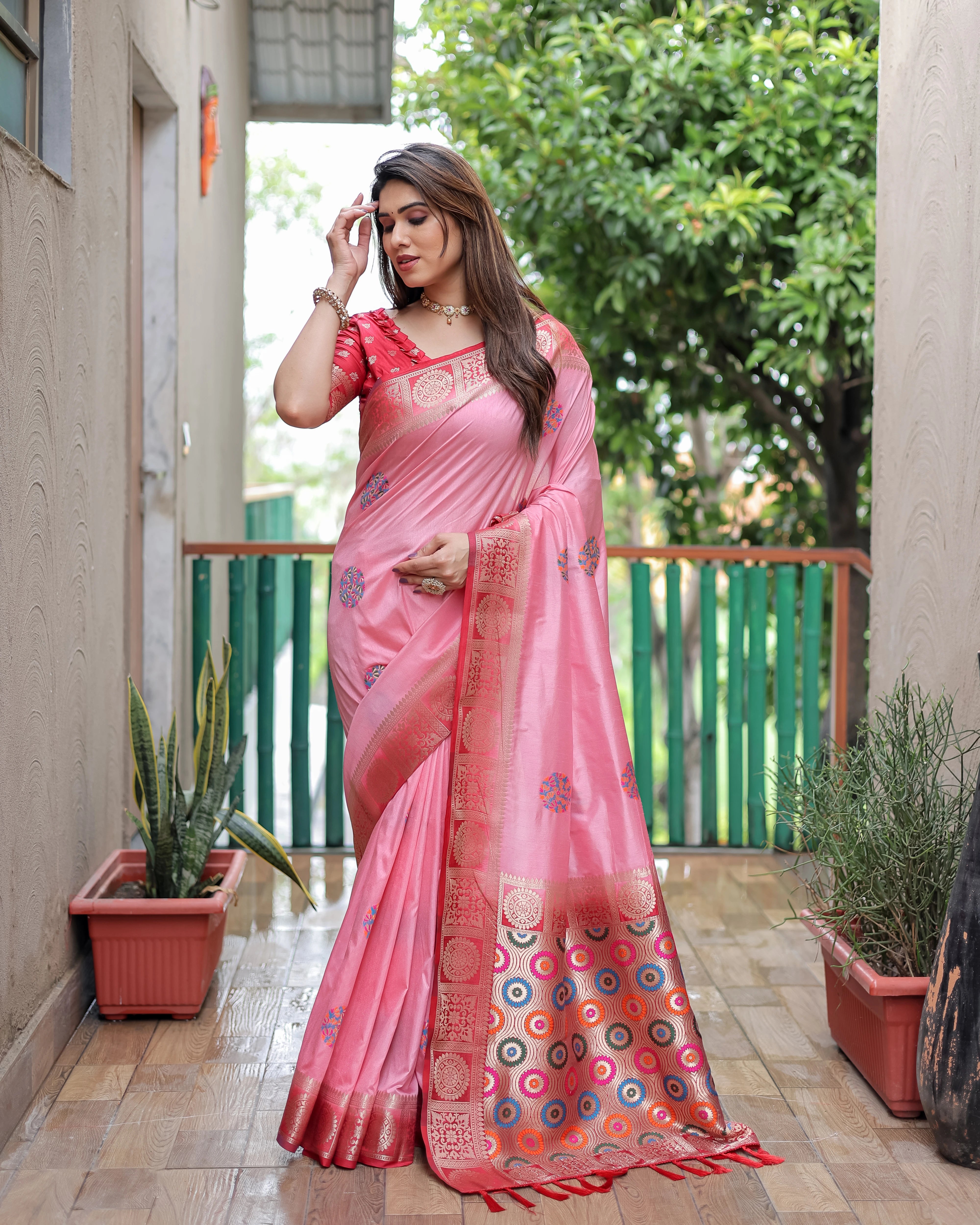 Woman in a pink saree standing outdoors with plants and a building in the background