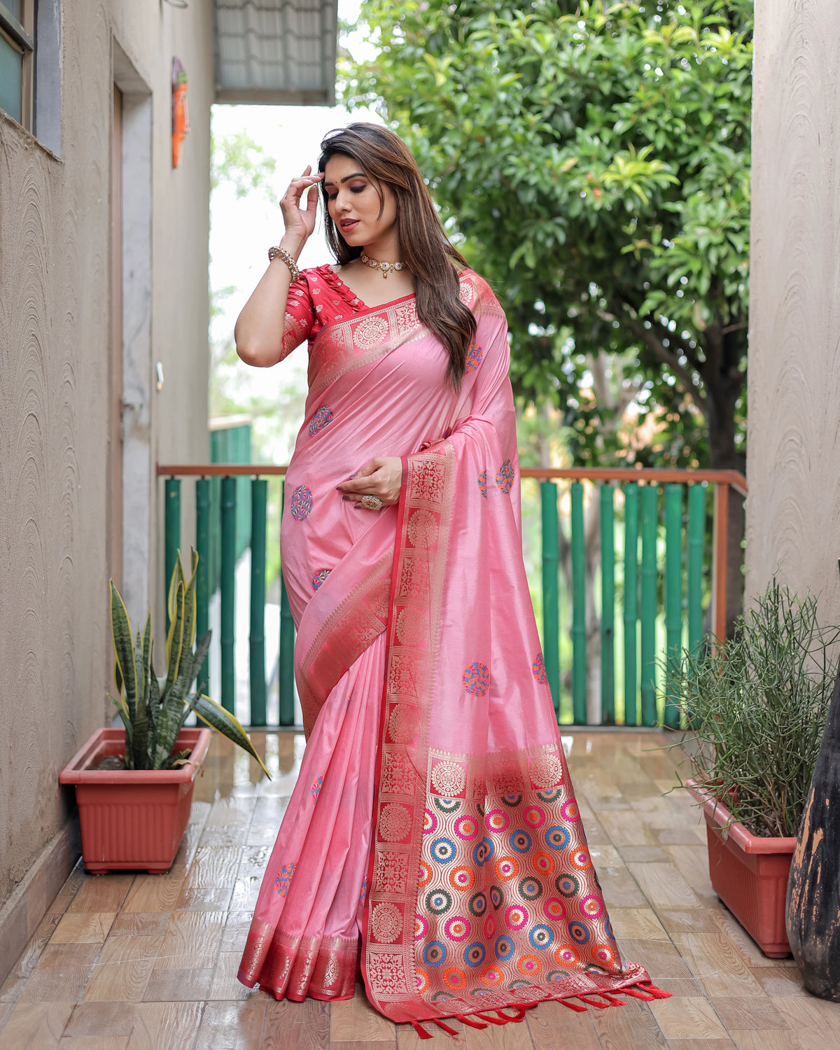 Woman in a pink saree standing outdoors with plants and a building in the background