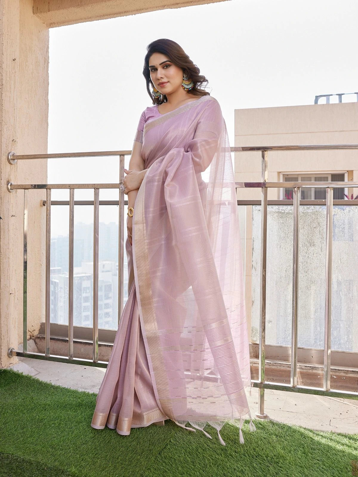Woman in a light pink saree standing on a balcony with a cityscape view.