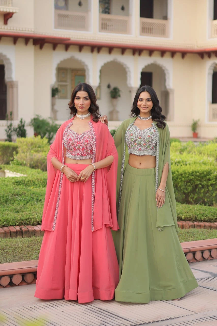 Two women in traditional pink and green outfits standing outdoors with a building and garden in the background.