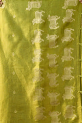 A close-up detail shot of the lime green saree fabric. The image highlights the texture of the material and the repeating woven silver cow motifs, characteristic of Pichwai-inspired designs. Small circular zari dots are scattered across the fabric, and green tassels are visible along the edge of the border.