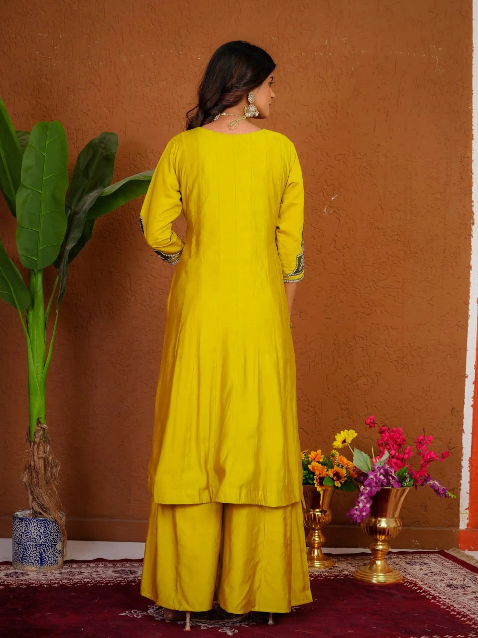 Woman in a yellow traditional outfit standing against an orange wall with decorative elements.