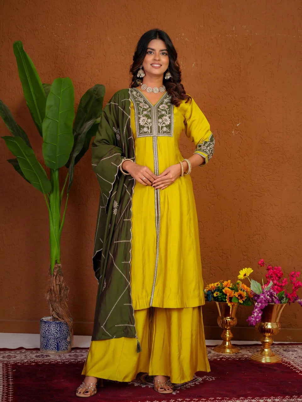 Woman in a yellow and green traditional outfit standing against a brown wall with decorative elements.