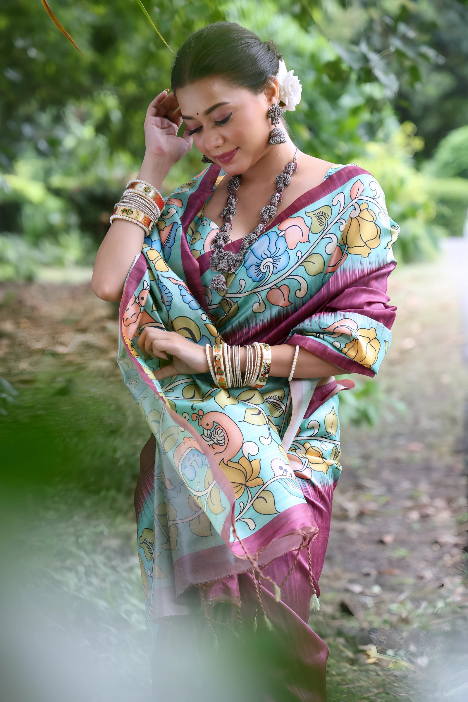 A woman models a stunning Magenta Soft Tussar Silk Saree. The pallu features a wide Sky Blue panel with an elaborate Kalamkari-style print of pink, yellow, and blue floral/peacock motifs. She wears a matching short-sleeve printed blouse and accessorizes with heavy oxidized silver jewelry, posing outdoors in a garden.