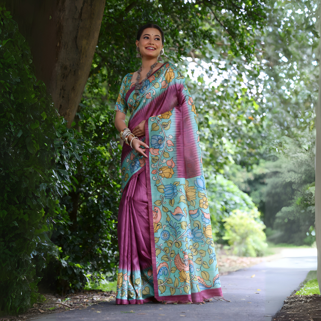 A woman models a stunning Magenta Soft Tussar Silk Saree. The pallu features a wide Sky Blue panel with an elaborate Kalamkari-style print of pink, yellow, and blue floral/peacock motifs. She wears a matching short-sleeve printed blouse and accessorizes with heavy oxidized silver jewelry, posing outdoors in a garden.