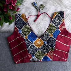  Multicolor Ajrak-print blouse with deep V-neck, featuring traditional geometric and floral patterns in blue, mustard, green, gold, and red, contrasted by solid red silk side panels with silver stripes, and tie-back detail, displayed flat on a white fur background with a bunch of artificial pink flowers in the corner.