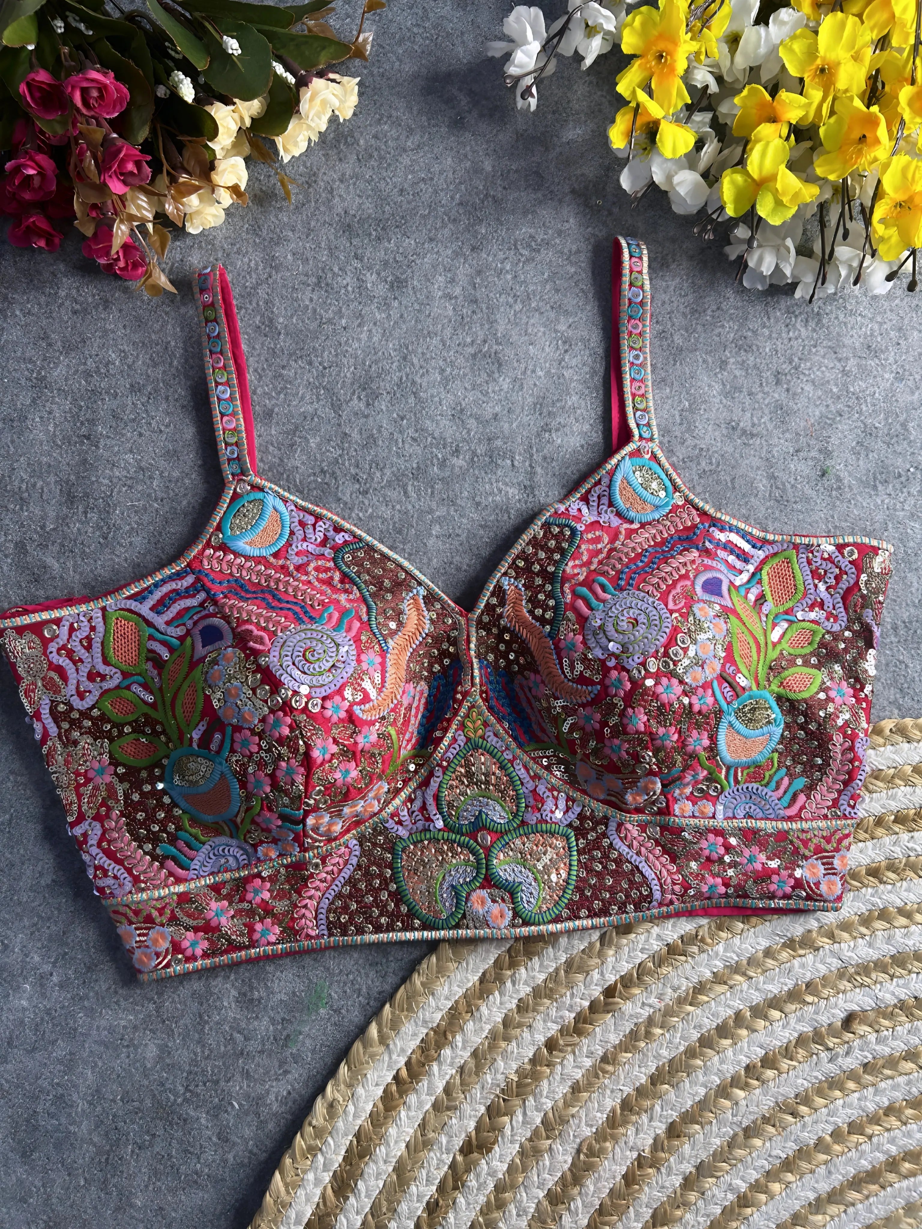Traditional multicolor bustier blouse featuring vibrant Madhubani-inspired embroidery with intricate floral, paisley, and geometric motifs, embellished with sequins and beads, structured cups, and adjustable straps, displayed flat on a textured grey surface with artificial pink, yellow, and white flowers in the corners.