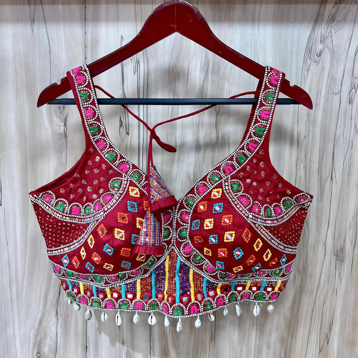 Traditional red Gujarati blouse featuring vibrant multicolor geometric embroidery, intricate mirror work, and white cowrie shell trim along the hem. The blouse has wide embroidered straps, structured cups, a decorative front center tassel, and tasseled tie-back detail, displayed on a wooden hanger against a light wood panel background.