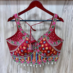 Traditional red Gujarati blouse featuring vibrant multicolor geometric embroidery, intricate mirror work, and white cowrie shell trim along the hem. The blouse has wide embroidered straps, structured cups, a decorative front center tassel, and tasseled tie-back detail, displayed on a wooden hanger against a light wood panel background.