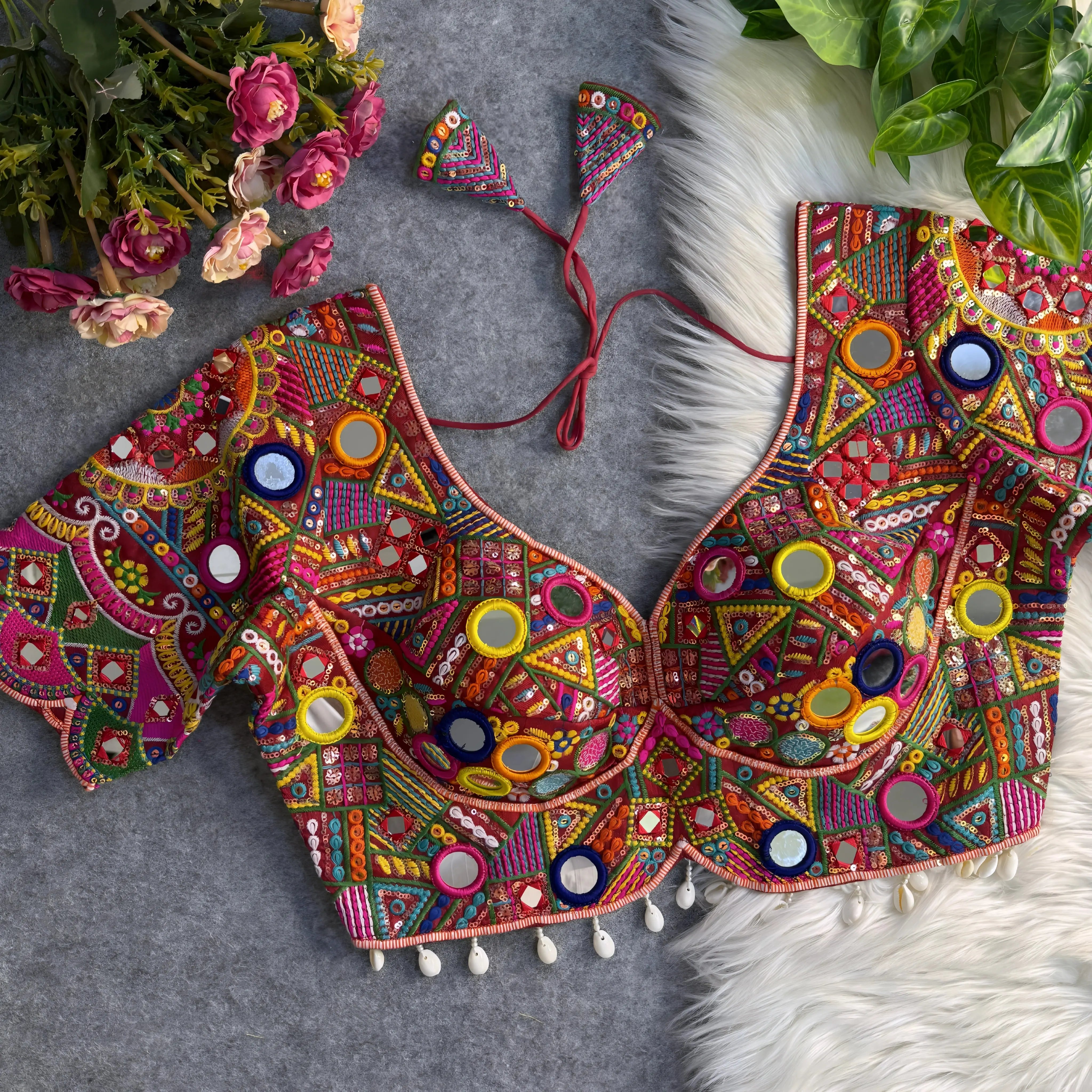 Traditional multicolor blouse featuring intricate Gujarati-style mirror embroidery with vibrant geometric and floral patterns in red, yellow, green, blue, pink, and orange. The blouse has short sleeves, a deep V-neckline with structured cups, shell-beaded trim along the hem, and tasseled tie-back detail, displayed flat on a textured grey surface with artificial pink flowers and green foliage.