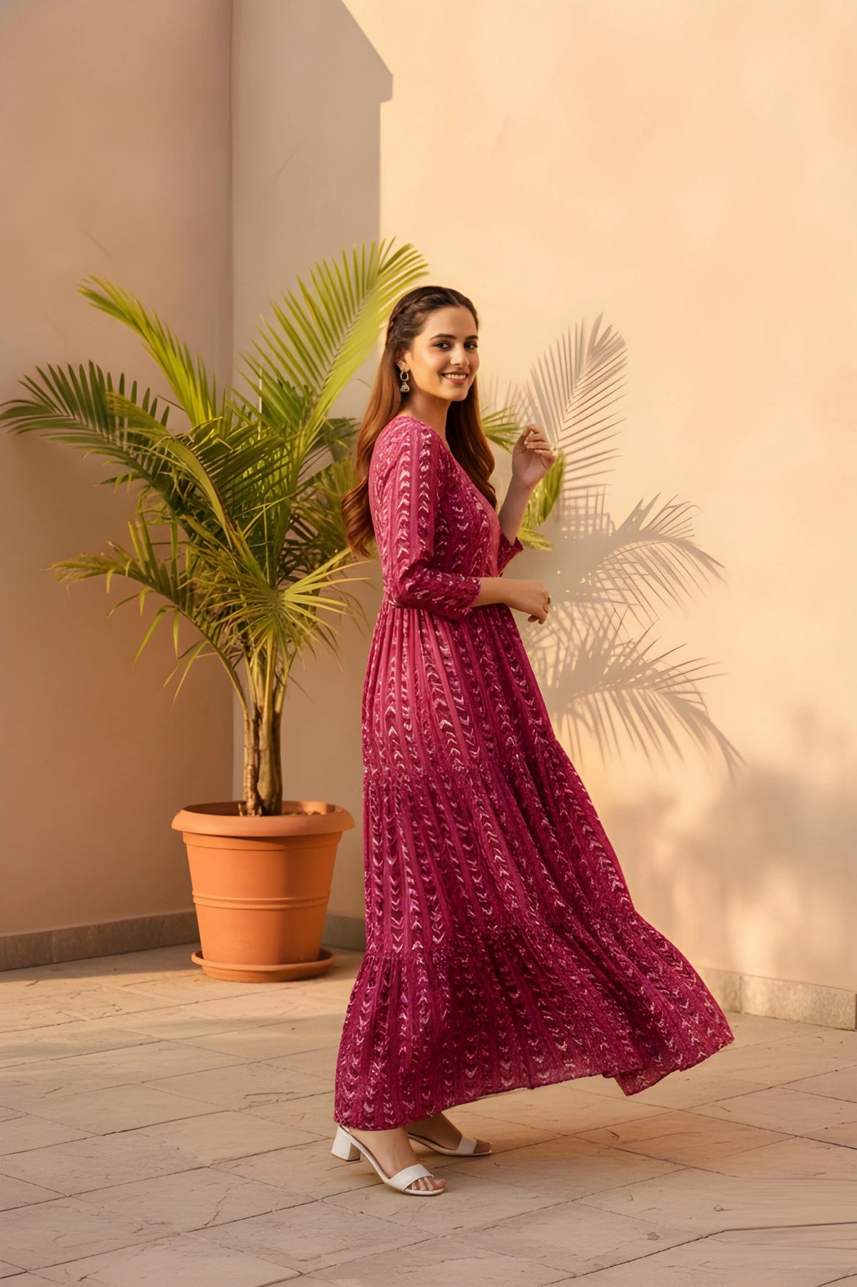 Woman in a red patterned dress standing next to a potted plant against a beige wall.
