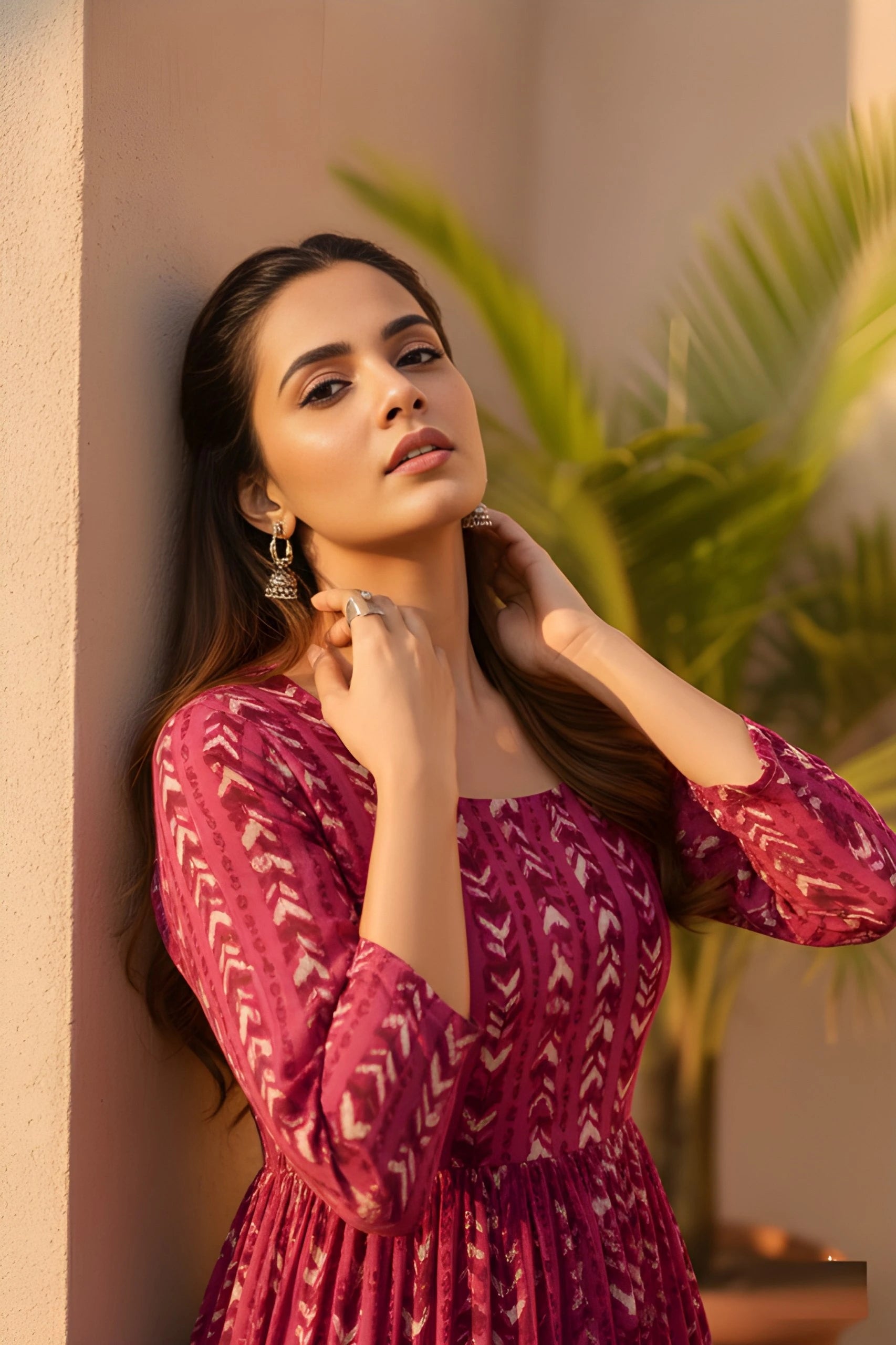 A close-up of a woman leaning against a wall in a magenta patterned dress, with her hands resting gracefully near her neck in warm sunlight.