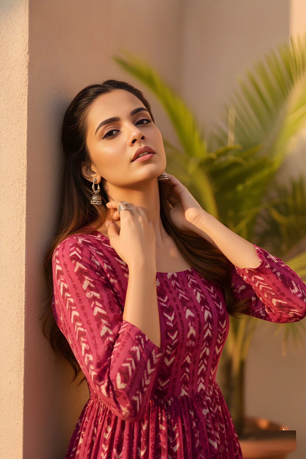 A close-up of a woman leaning against a wall in a magenta patterned dress, with her hands resting gracefully near her neck in warm sunlight.