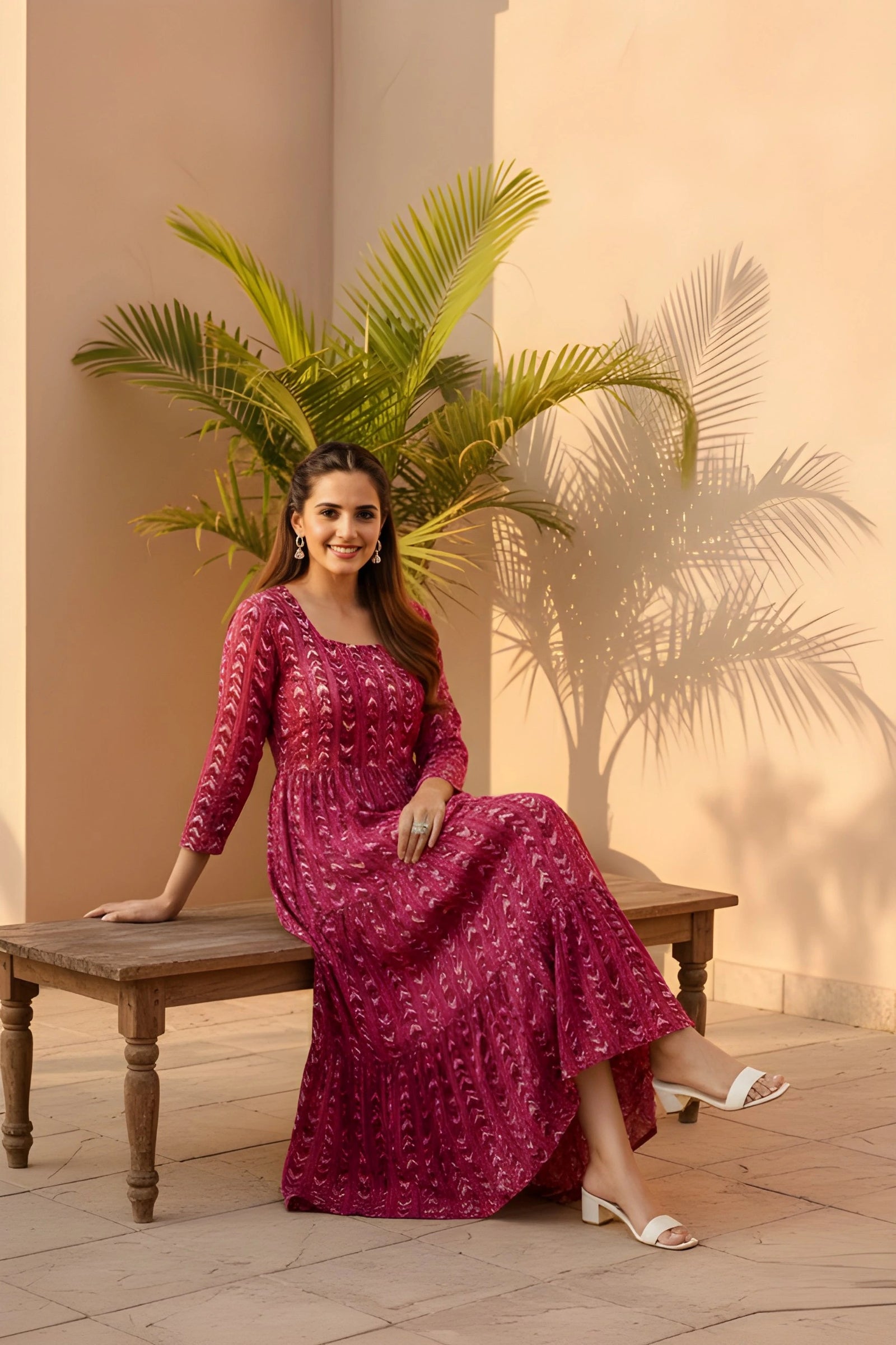 A woman sitting on a rustic wooden bench outdoors, wearing a magenta patterned dress and white sandals, smiling at the camera.