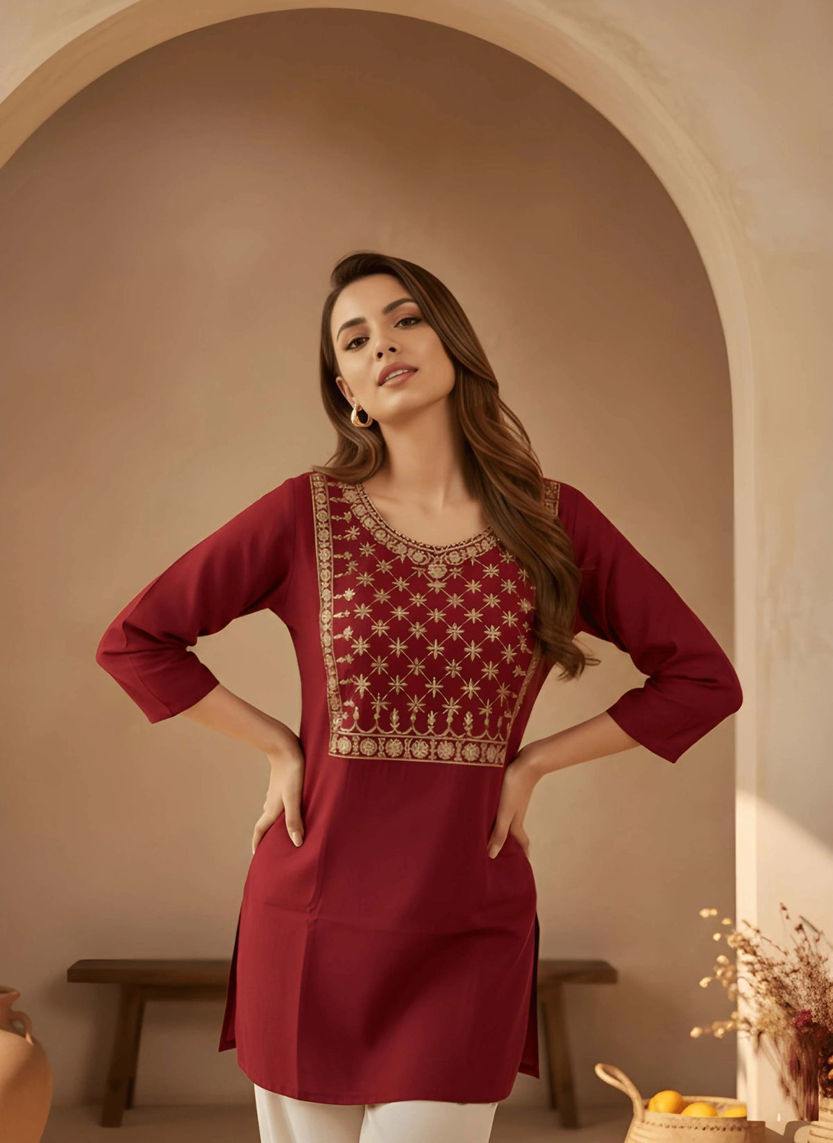 Woman wearing a maroon traditional outfit with intricate patterns in a warm-toned room.