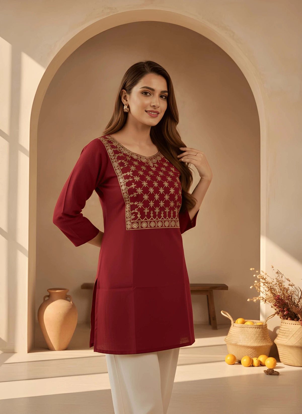 Woman wearing a maroon traditional outfit with intricate gold embroidery in a warm-toned interior setting.