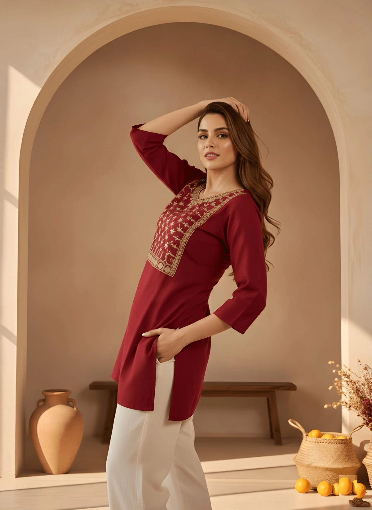 Woman wearing a red embroidered top and white pants in a warm-toned interior setting.
