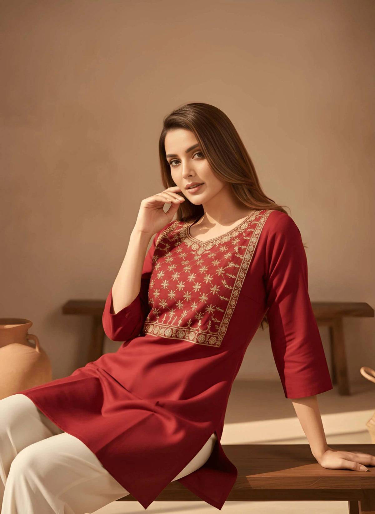 Woman wearing a red traditional outfit with gold embroidery, sitting on a wooden stool against a beige wall.