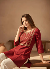 Woman wearing a red traditional outfit with gold embroidery, sitting on a wooden stool against a beige wall.