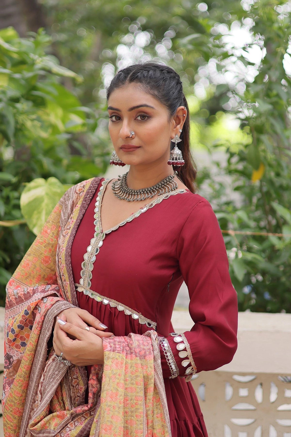 Woman in traditional attire with a red blouse and patterned dupatta, standing outdoors with greenery.