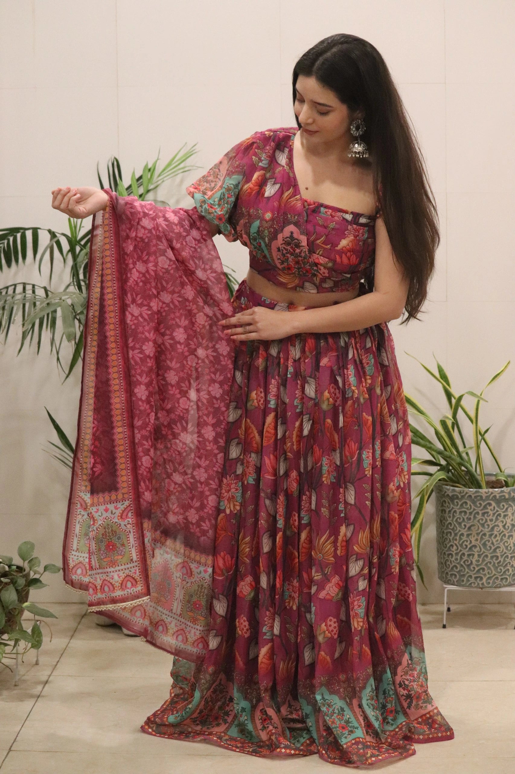 Woman in a floral saree with a matching dupatta, standing indoors with plants in the background.