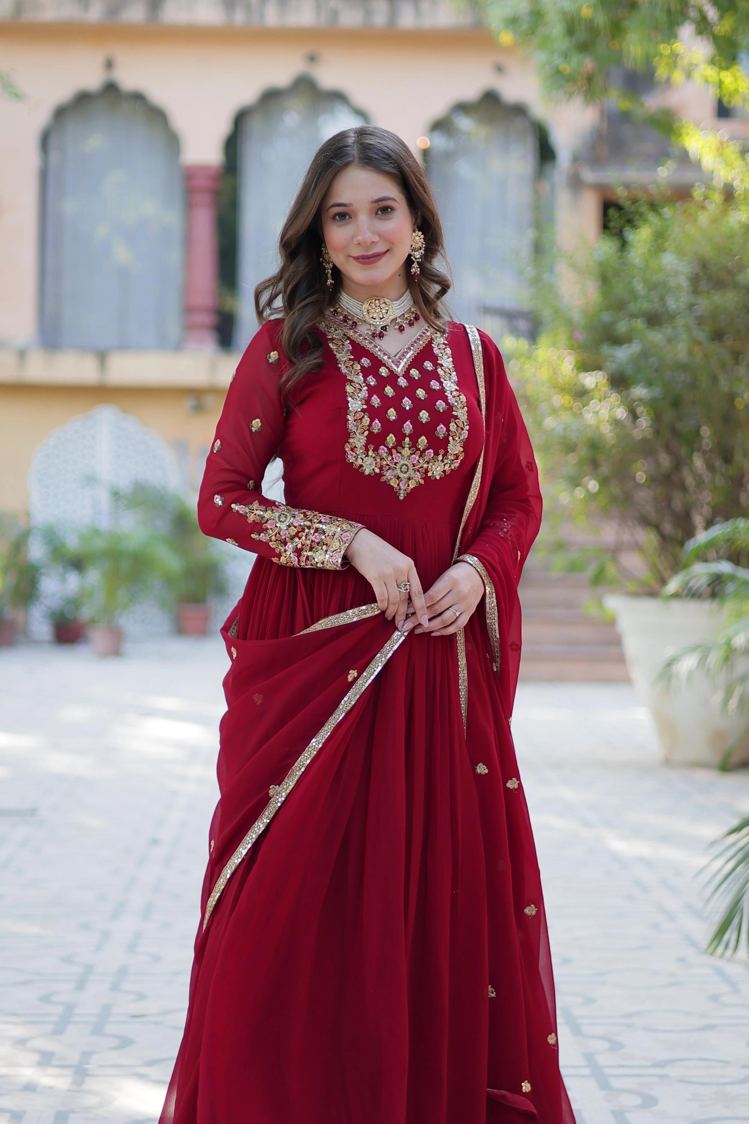 A full-length front view of a woman in a red ethnic gown with gold embroidery. She is looking toward the camera with a gentle smile, wearing a pearl and gold choker necklace. The background shows a building with traditional arched windows and greenery.